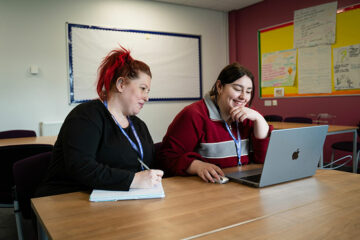 Two people sat together at a table both looking at the same laptop