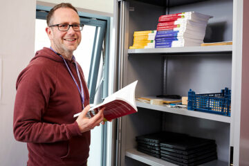 A person stood with an open book with shelves of books next to them