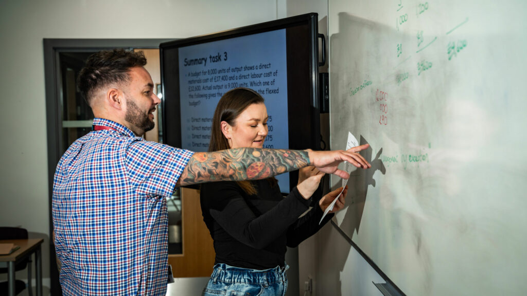 A student writing on a white board and a lecturer stood pointing at the white board