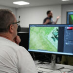 An adult sat in front of a computer with the lecturer in the background