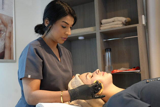 A lady with black hair, grey uniform and black gloves with her hands on a clients face who's laying on a treatment bed