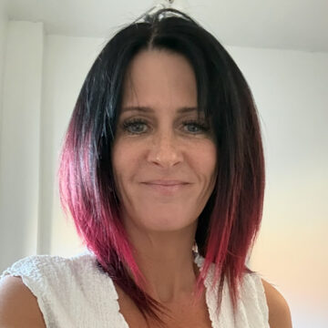 A head and shoulders photo of a smiing lady with black and red hair and a white top