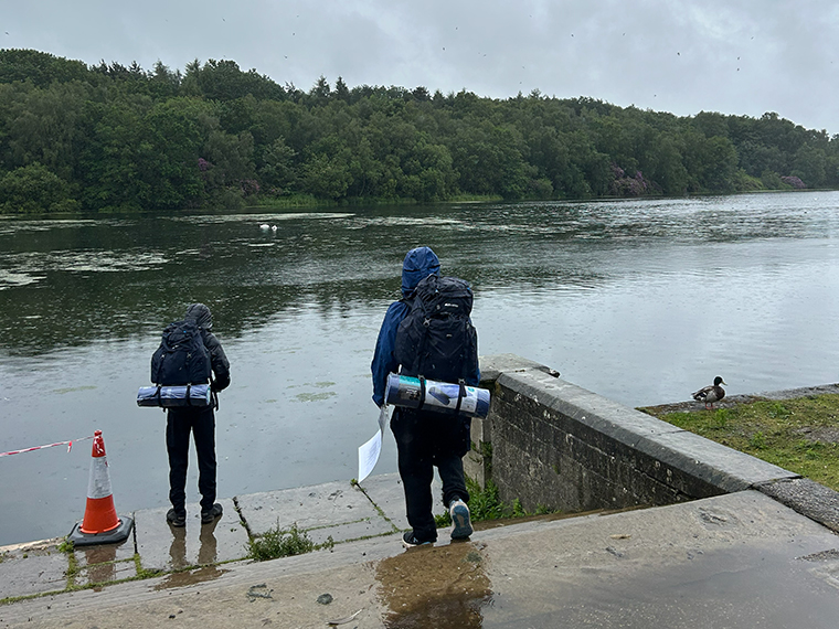 Two people wearing coats and backpacks walking toward a lake, a line of trees are across the other side of the lake