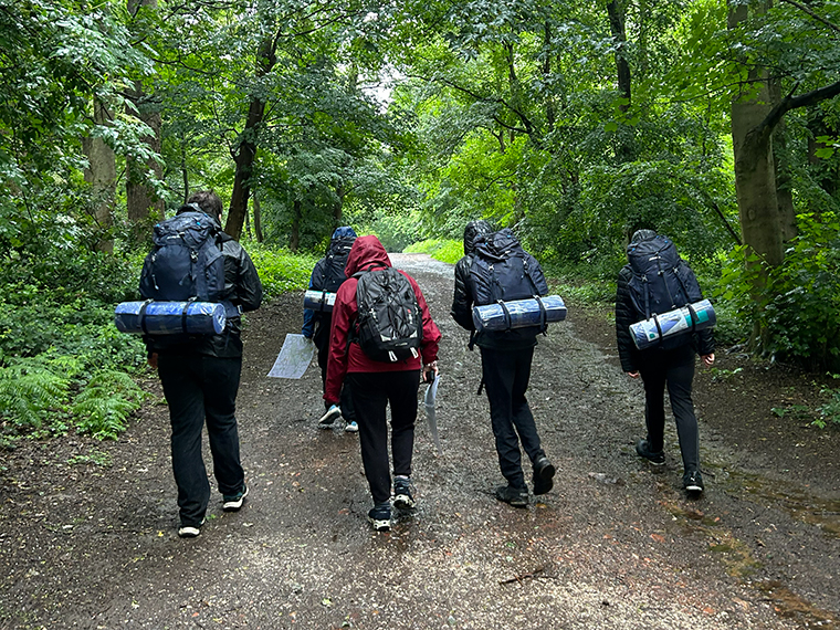 Five people wearing coats and holding backpacks, walking up a path with trees either side
