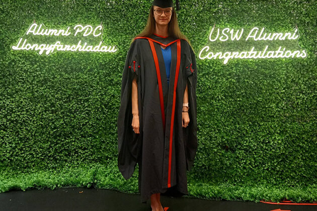 Hannah stood in her graduation attire in front of a green hedge with lit up words 'USW Alumni Congratulations'
