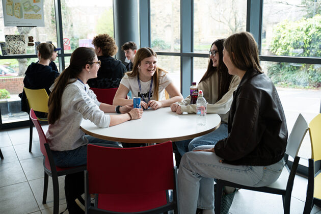 Two tables each full of smiling students socialising, large windows behind the tables