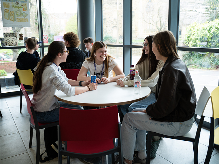 Two tables each full of smiling students socialising, large windows behind the tables