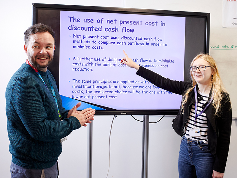 Accounting tutor Conal stood at a large TV screen supporting a student who is presenting and pointing to the screen